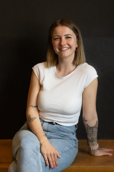 Eine Frau mit schulterlangem Haar, weißem Top und hellblauen Jeans sitzt lächelnd auf einer Holzbank.